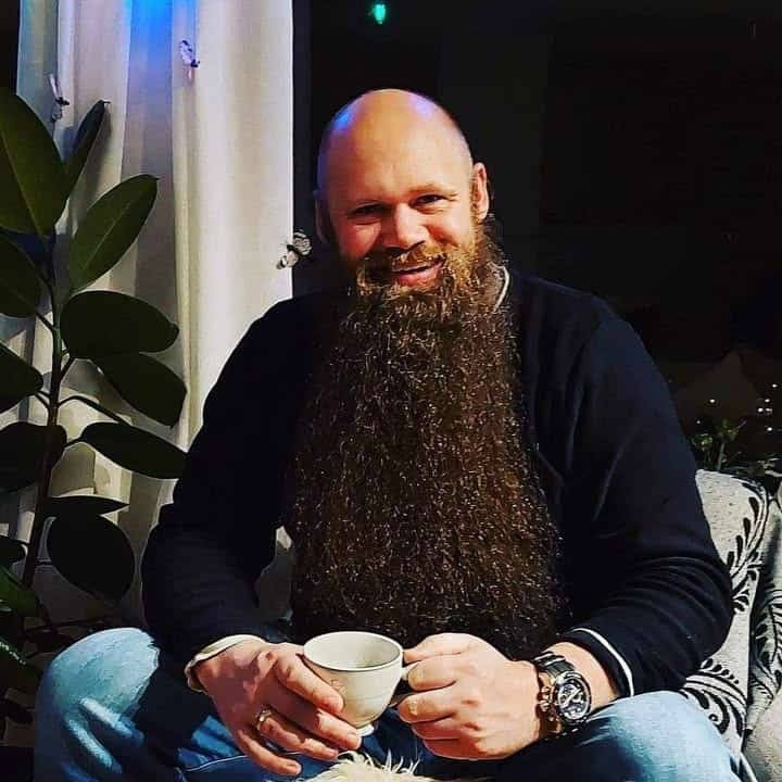 Smilende mann med gigantisk skjegg sitter med kaffekopp og stålur ved siden av en plante.